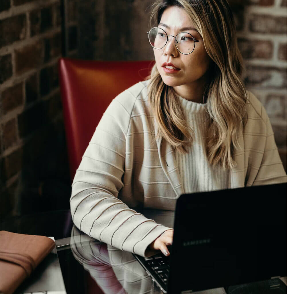 'http://Woman%20sitting%20in%20a%20red%20chair%20and%20glass%20table%20working%20on%20her%20laptop%20looking%20out%20a%20window